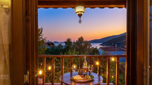 A wooden balcony with a table set for dining, overlooking a scenic view of the sea and hills at dusk.