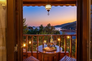 Un balcón de madera con una mesa lista para comer, con vistas a un paisaje marino y montañoso al anochecer.