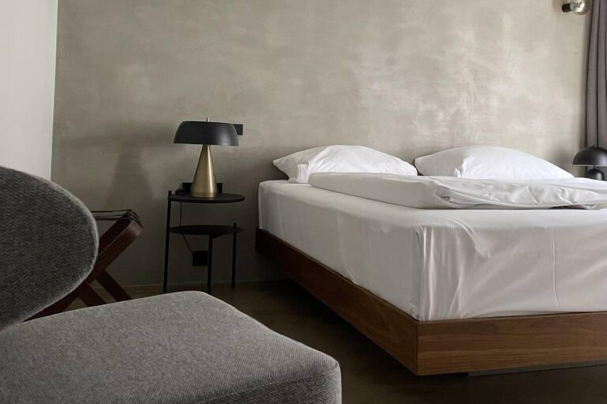 A modern hotel room with a single bed, a small bedside table with a lamp, and a grey armchair.