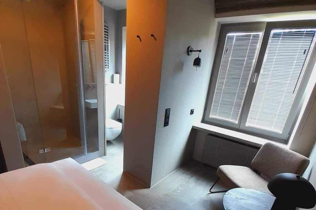 A modern hotel room with a bed, a small round table, a chair, a window with blinds, and a bathroom visible through an open door.