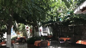Un café en plein air avec des tables et des chaises disposées sous des arbres, des murs en pierre et un sol carrelé.