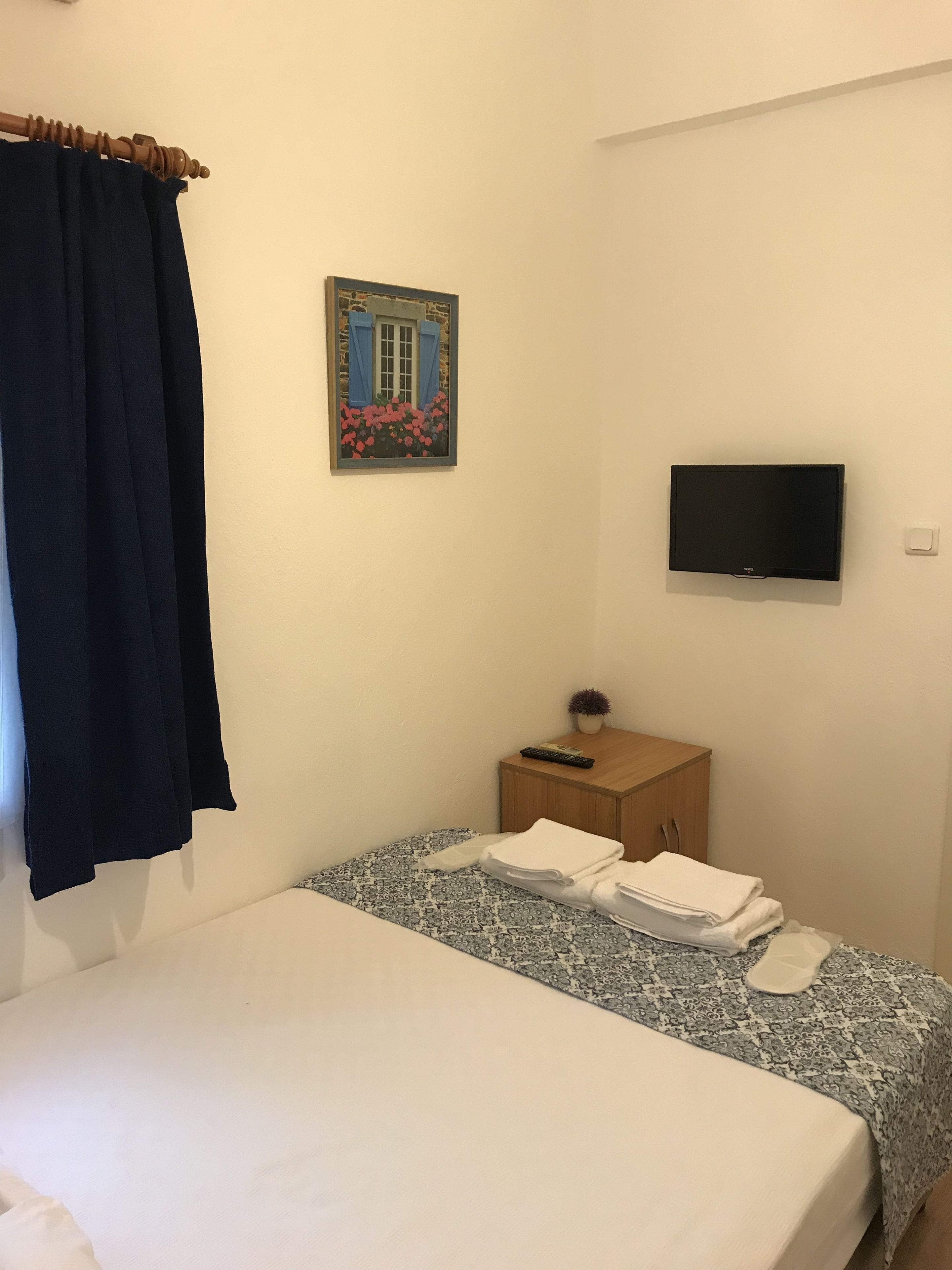 Ett sovrum med en säng, en tv som är monterad på väggen, ett litet träbord vid sängen med en fjärrkontroll och en tavla som föreställer ett hus.