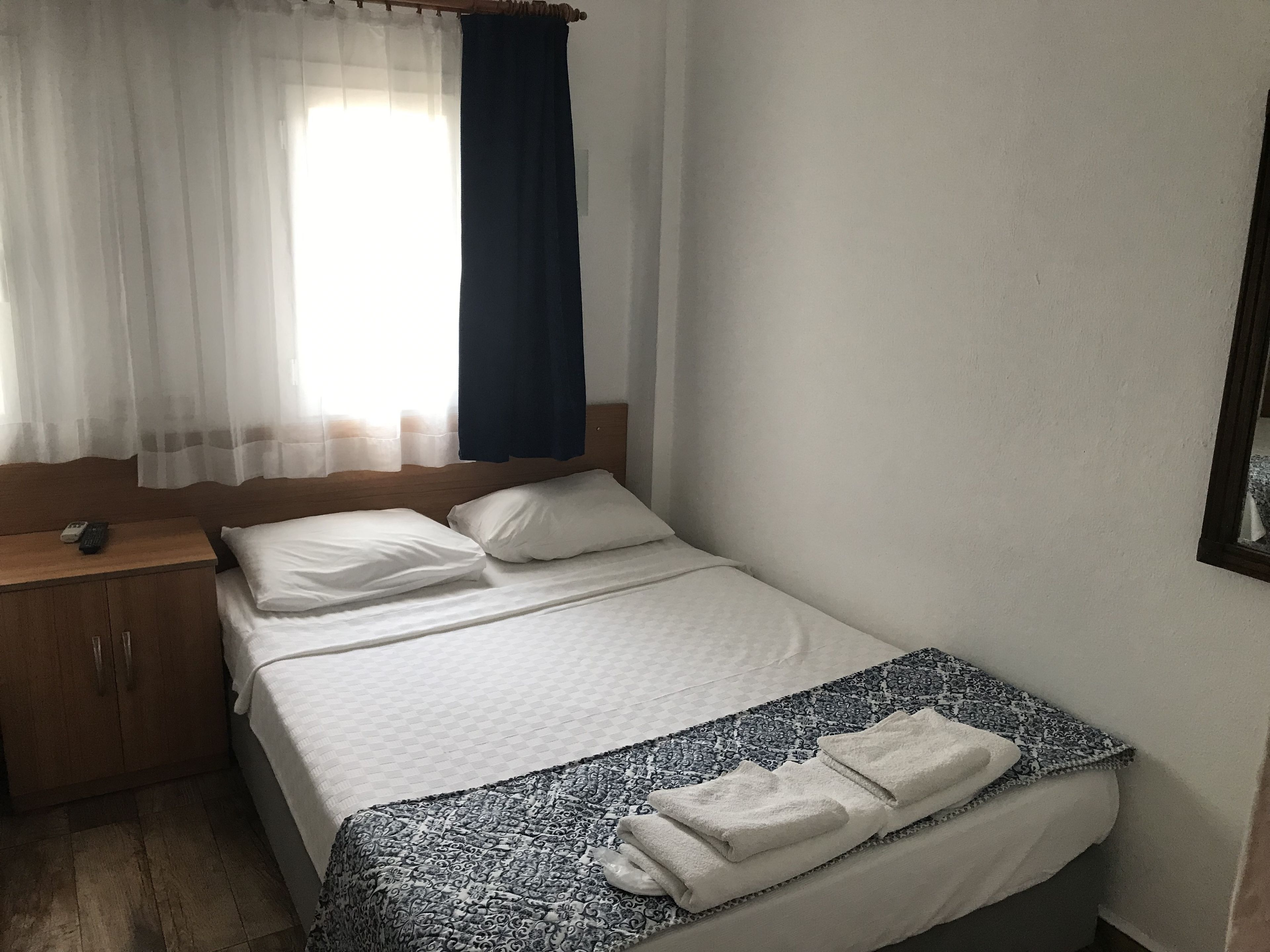 Ein Einzelbett mit weißen Laken, ein Nachttisch aus Holz mit Fernbedienung, ein dunkelblauer Vorhang und eine dekorative Decke mit Muster.