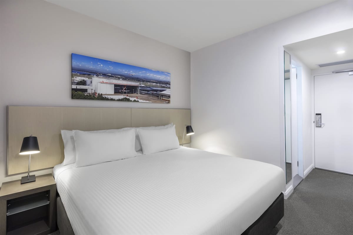 A modern hotel room with a large bed, a nightstand with a lamp, a painting on the wall, and a door leading to another room.