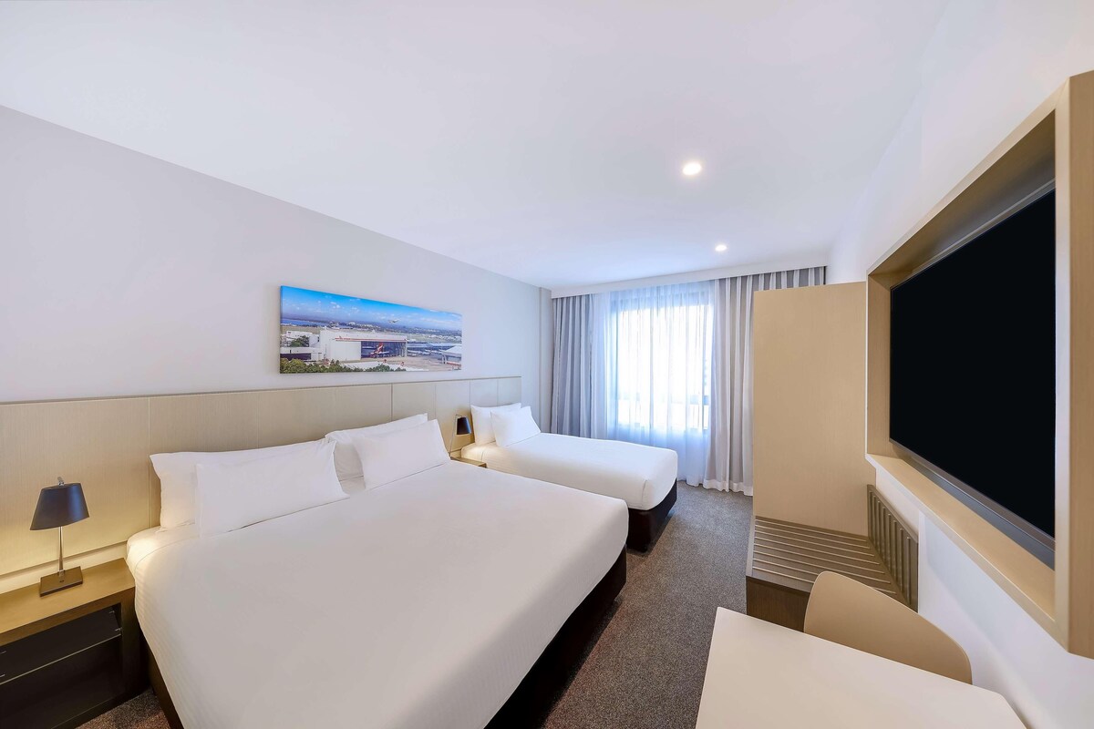 A hotel room with two beds, a flat-screen TV, a painting on the wall, and a window with curtains.