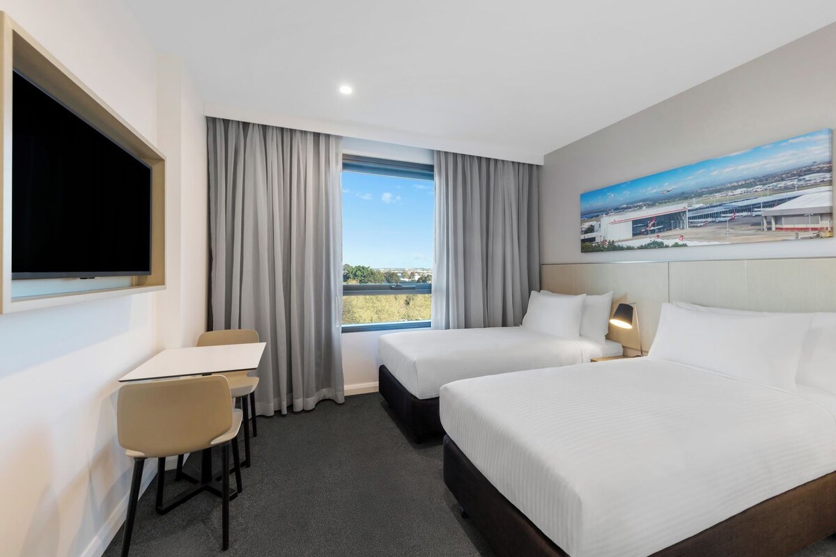 A hotel room with two beds, a desk, a chair, and a large window with curtains.