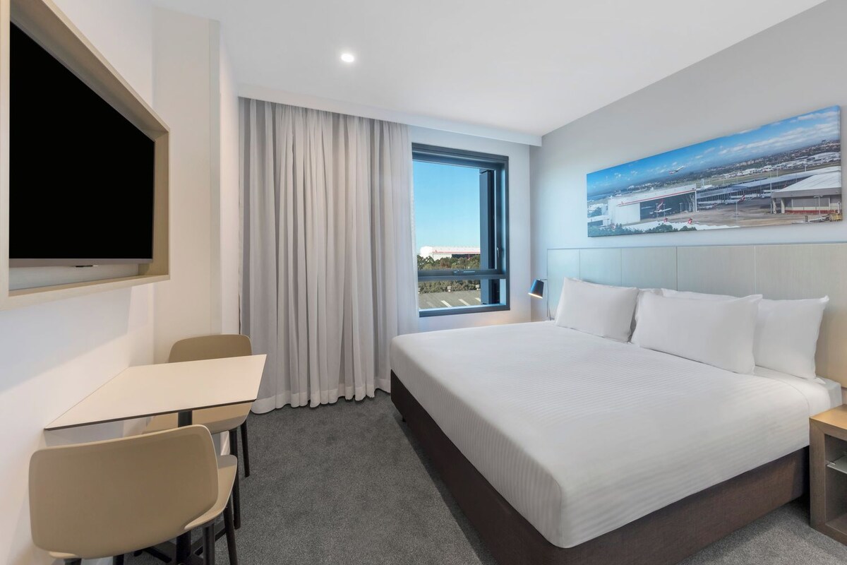 A hotel room with a bed, a desk, chairs, a TV, a window with curtains, and a painting on the wall.
