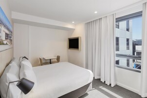 A hotel room with a bed, a desk, a chair, a TV, and a large window with curtains.