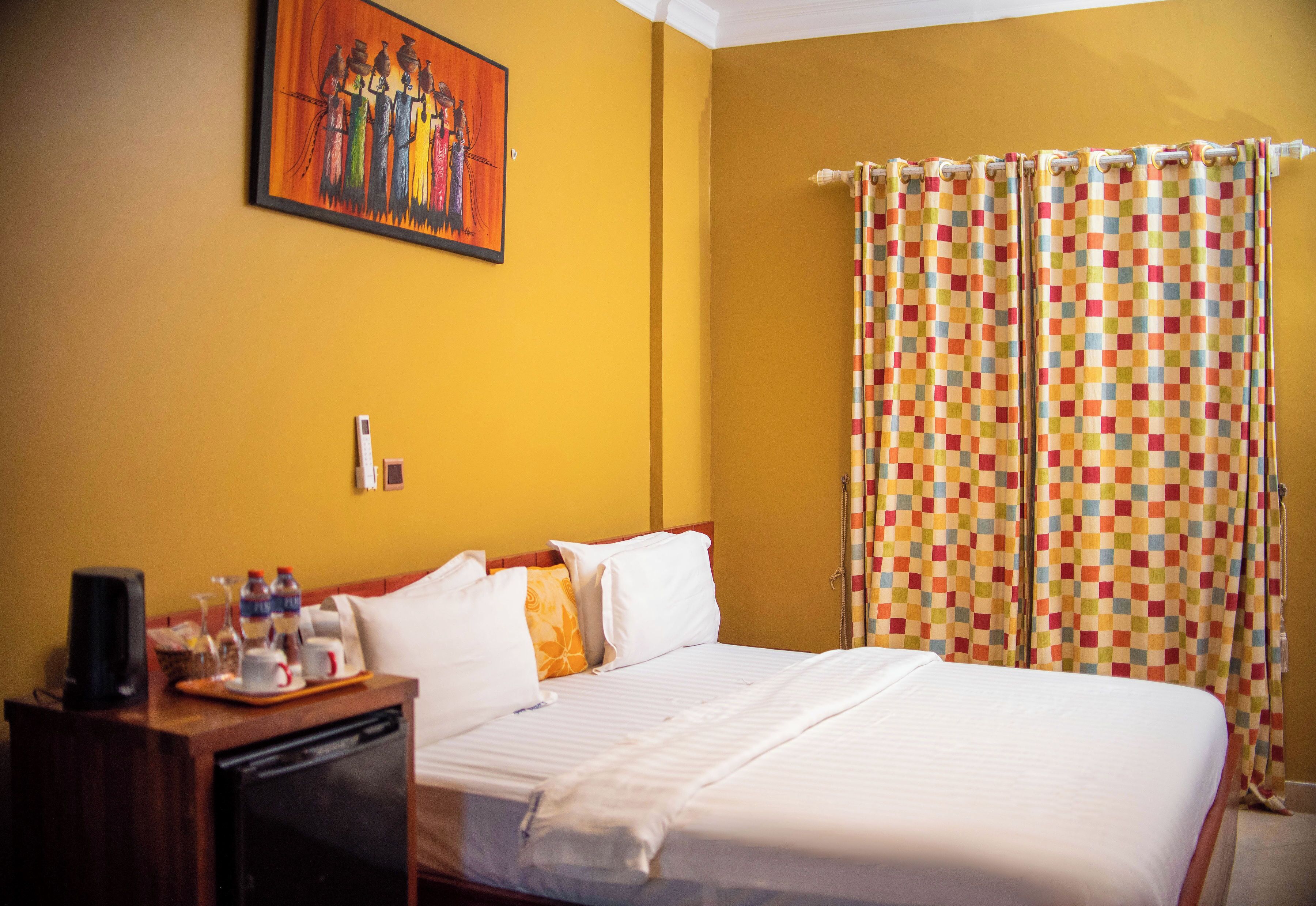 Habitación de hotel con una cama, una mesita de noche con cafetera, una bandeja con tazas y botellas, y una cortina colorida a cuadros.