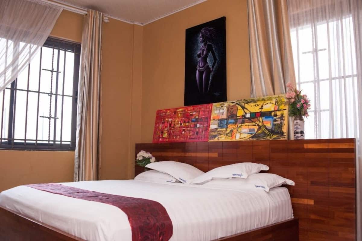 A hotel room with a wooden headboard, a large bed with white linens, a colorful abstract painting above the bed, and a window with curtains.