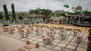 An outdoor café with multiple tables and chairs, surrounded by potted plants and a stage area. - Triple Hill Hotel (Janman)