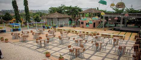 Un café en plein air avec de nombreuses tables et chaises, entouré de plantes en pot et doté d’une petite scène.