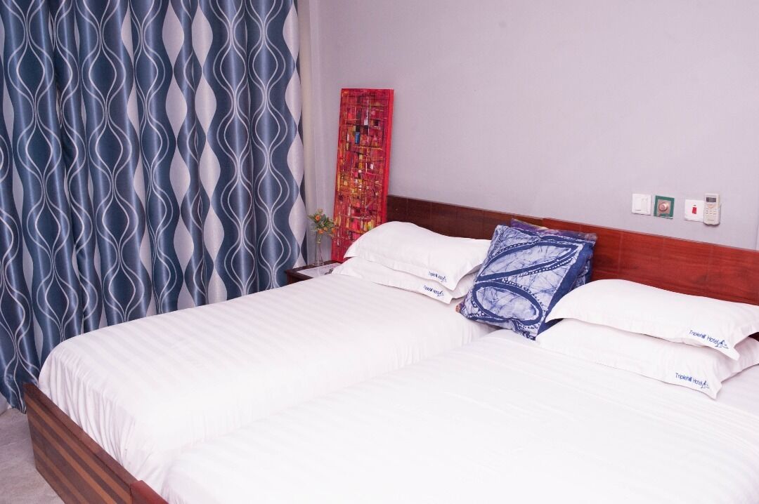 Een hotelkamer met een bed, wit beddengoed, een blauw-wit gestreept gordijn, een rode afbeelding aan de muur, en een houten hoofdeinde.