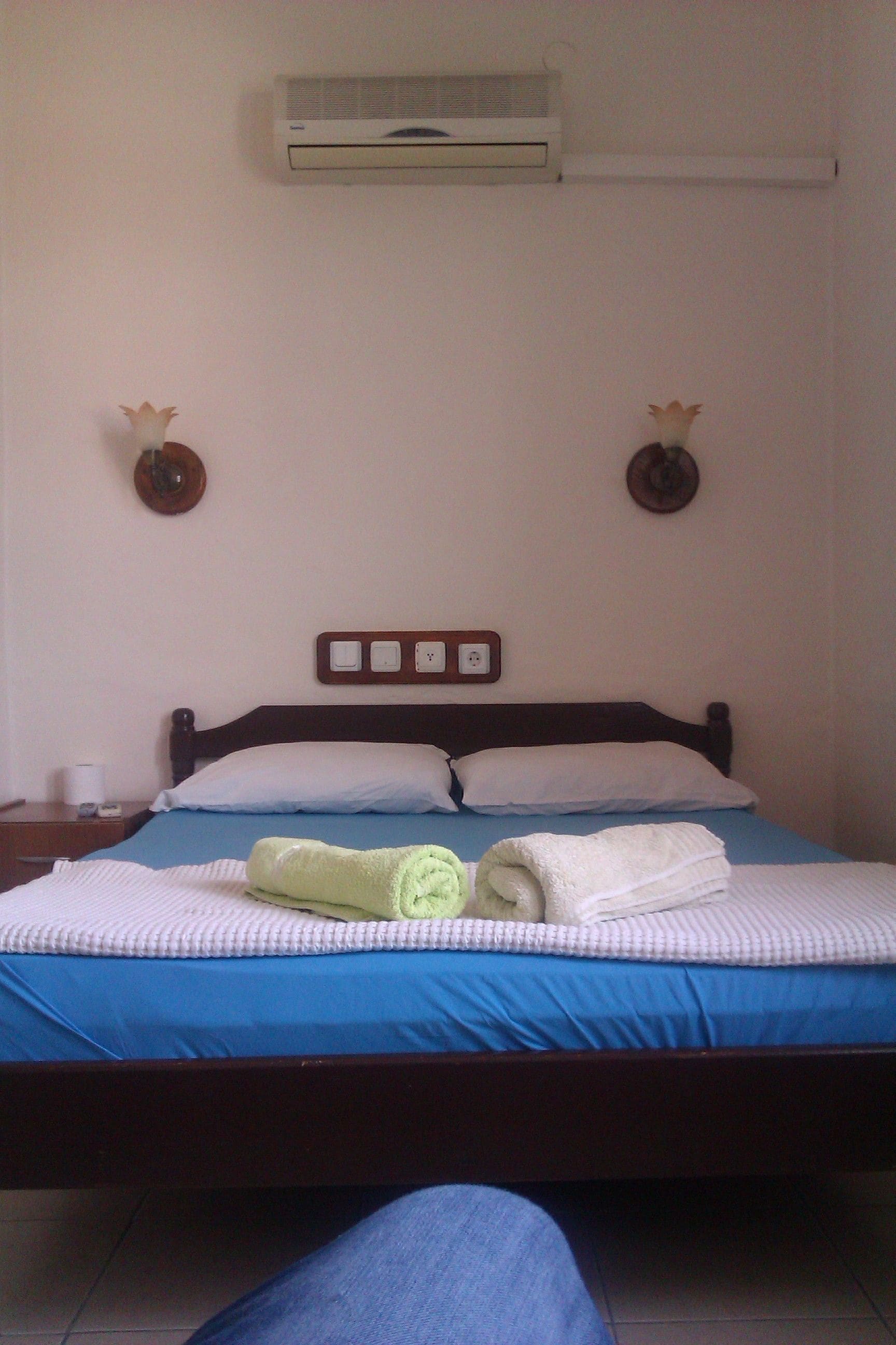 A bedroom with a bed, an air conditioner, a nightstand, and two wall-mounted lamps.