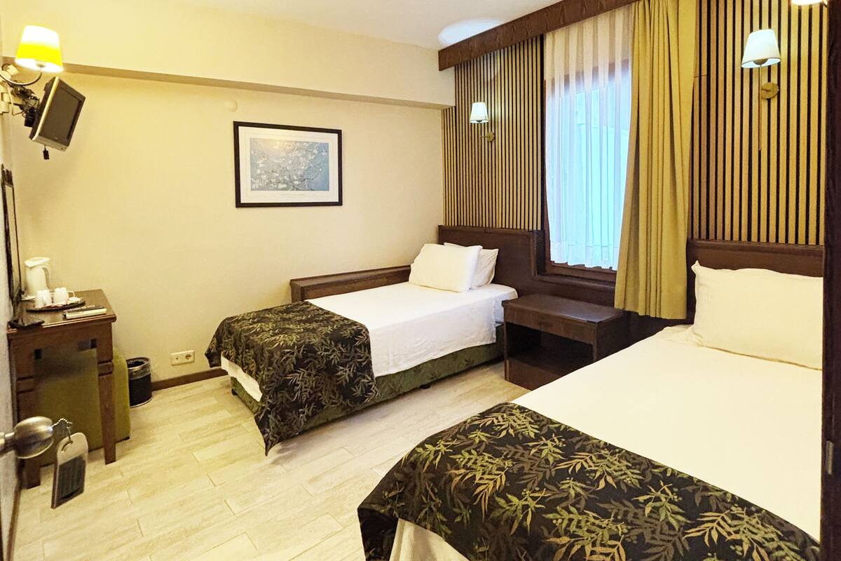 A hotel room with two beds, a desk with a lamp, a picture on the wall, and a window with curtains.