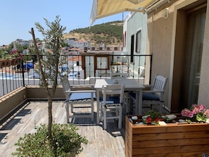 A terrace with a table and chairs, a wooden planter with flowers, and a view of the surrounding buildings and hillside. - Mitos Hotel (Bozcaada)