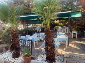 An outdoor seating area with tables and chairs, surrounded by palm trees and potted plants. - Mitos Hotel (Bozcaada)