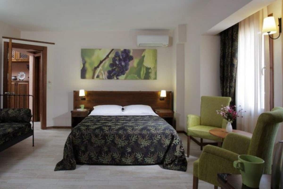 A hotel room with a bed, a sofa, a chair, a table with a cup, and a painting on the wall.