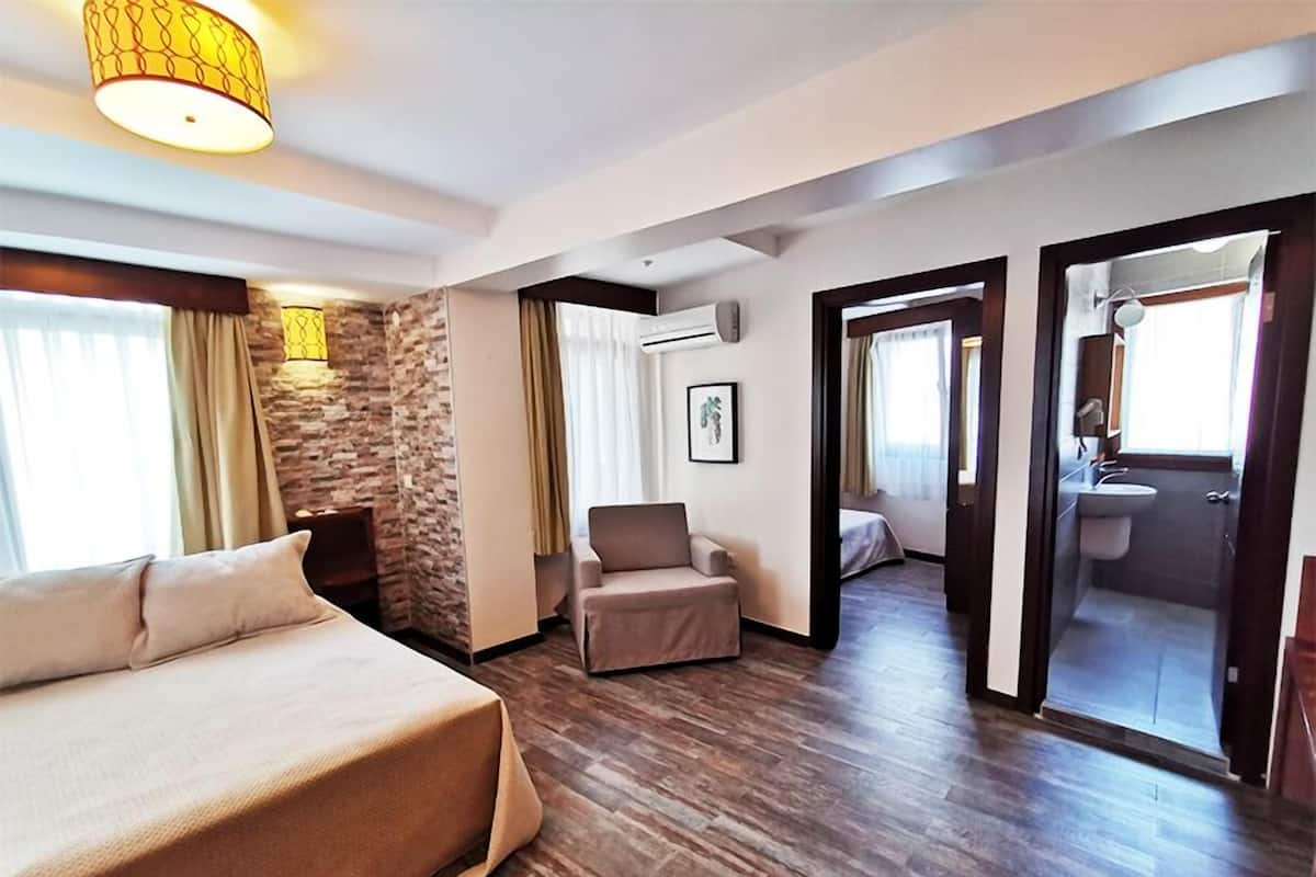 A hotel room with a large bed, a seating area, a stone accent wall, and a bathroom visible through an open door.
