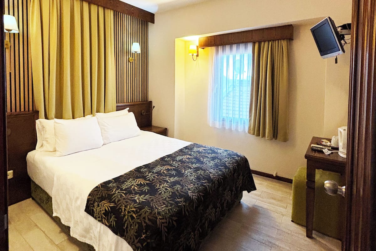 A hotel room with a bed, curtains, a desk with a monitor, and a window with blinds.