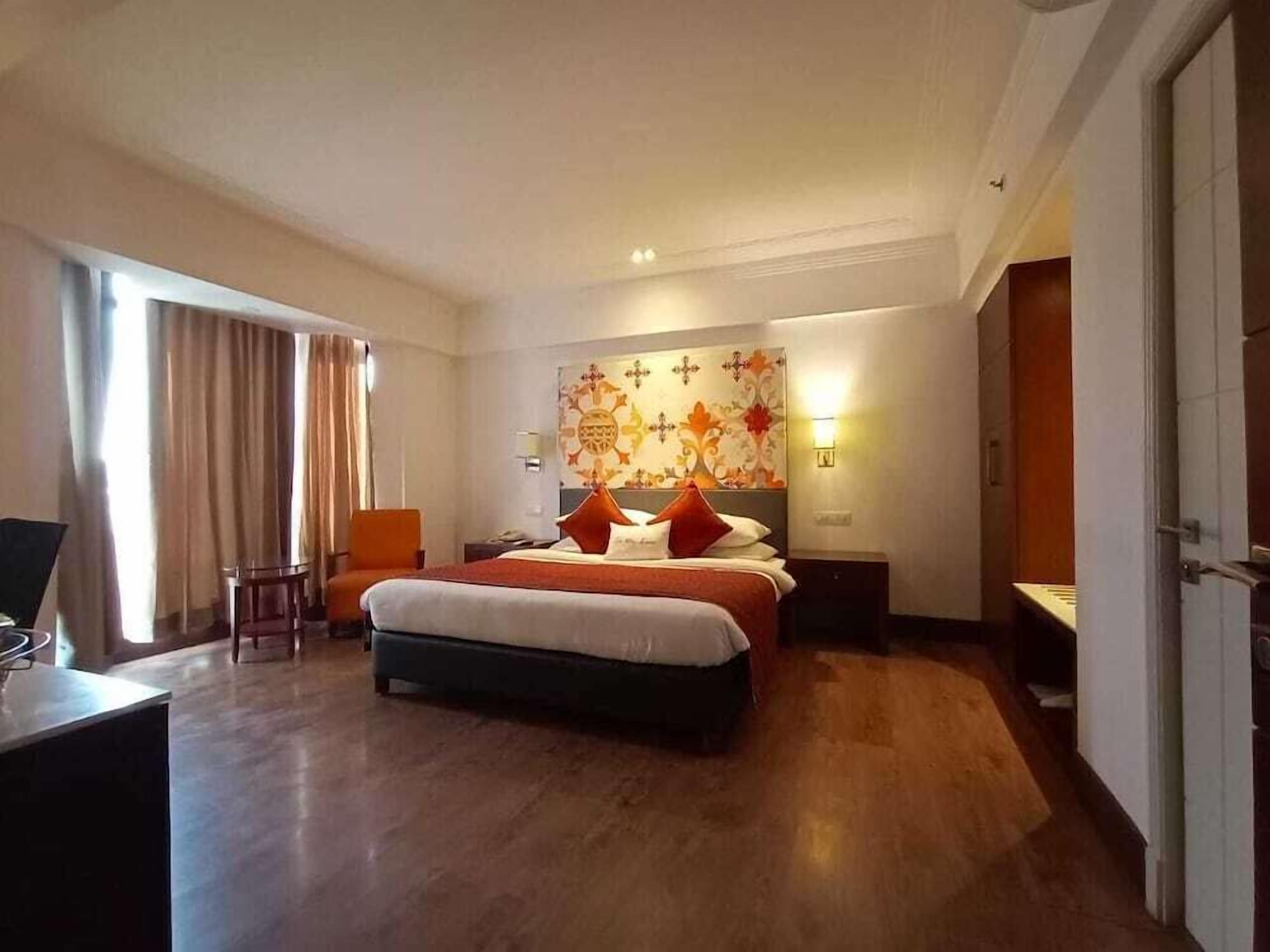 A hotel room with a bed, a desk, a chair, a nightstand, and a wall with a decorative pattern.
