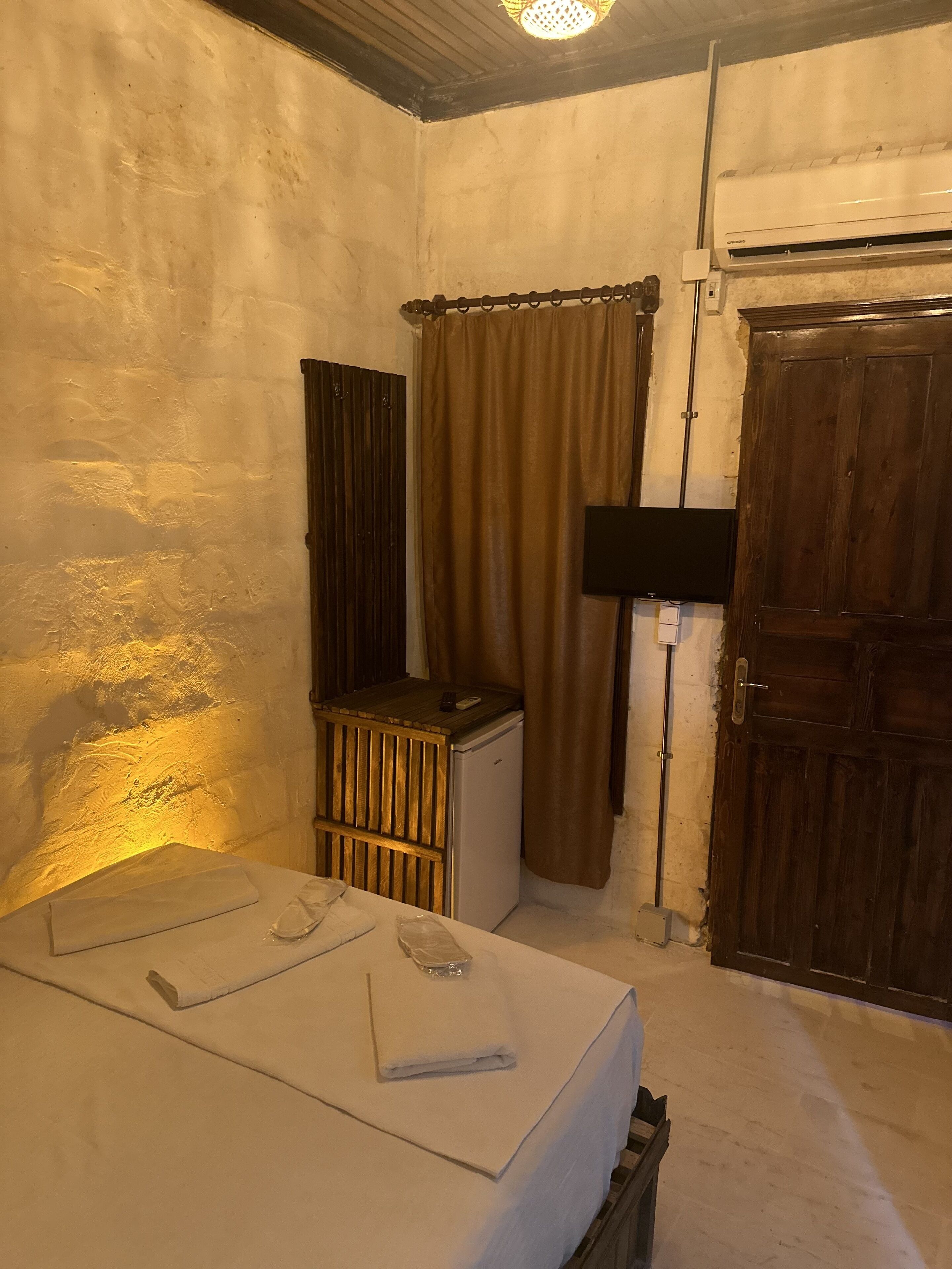 A small, dimly lit room with a single bed, a small heater, a window with curtains, a wooden door, and a wall-mounted air conditioning unit.