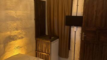 A small, dimly lit room with a single bed, a small heater, a window with curtains, a wooden door, and a wall-mounted air conditioning unit.