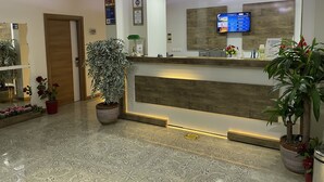 A hotel reception area with a wooden reception desk, a digital screen displaying information, and several potted plants. - Hotel Santana Life (Edremit)