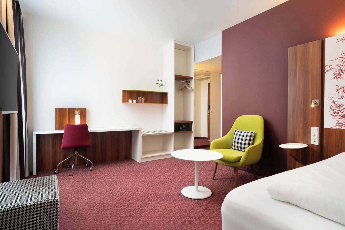 Una habitación de hotel con un escritorio, un sillón amarillo, un sofá blanco, un otoman estampado y una alfombra roja.
