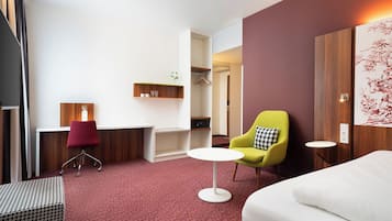 A hotel room with a desk, a yellow armchair, a white sofa, a patterned ottoman, and a red carpet.