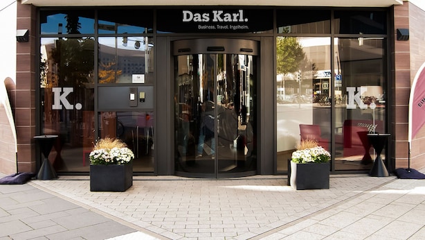 Ein Gebäudeeingang mit Glasfassade, einem Schild mit der Aufschrift "Das Karl" und zwei Blumenkästen am Bürgersteig.