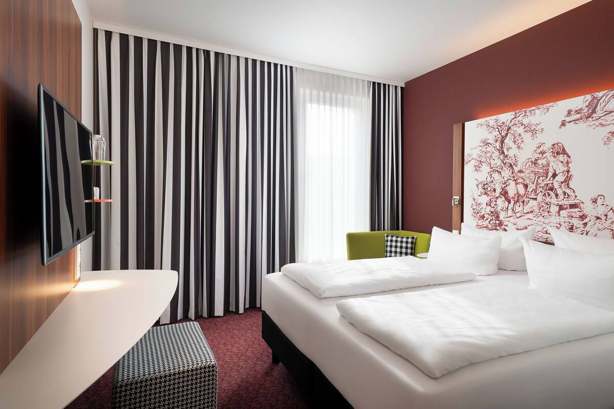 Habitación de hotel con cama, televisión montada en la pared, escritorio y ventana con cortinas a rayas.