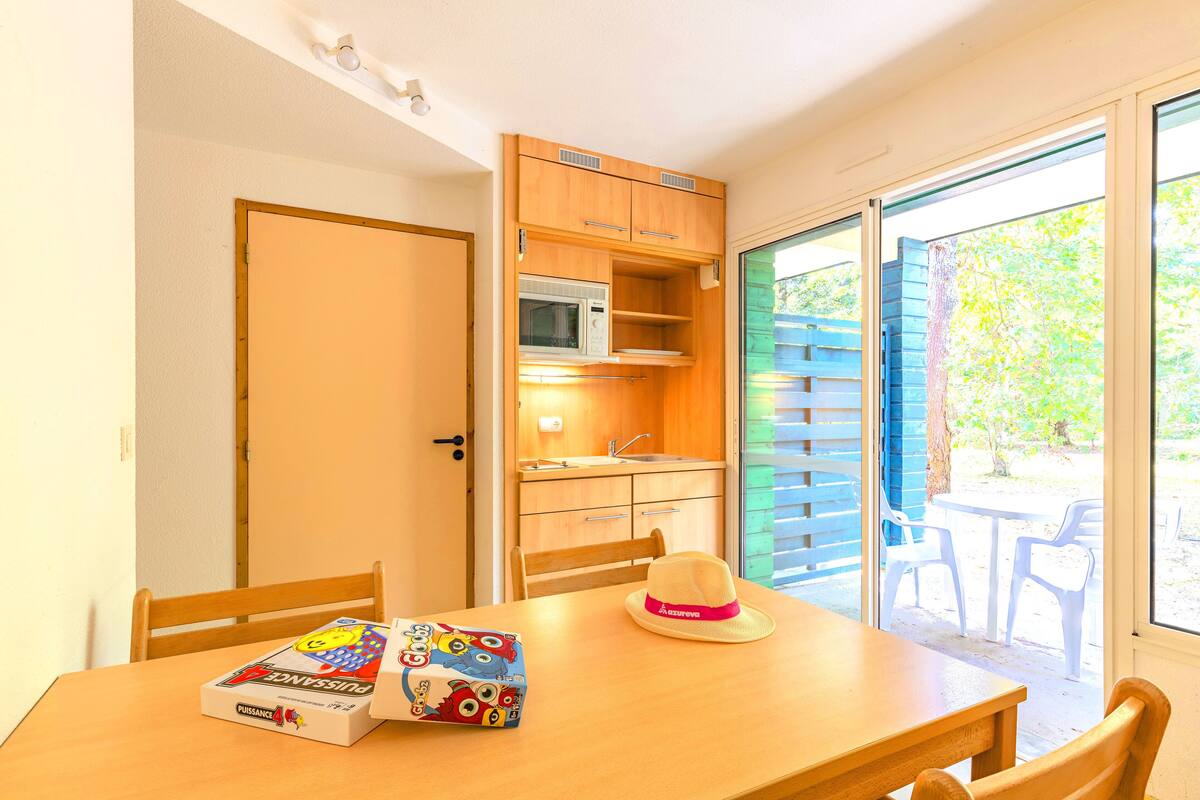 A compact kitchen with a microwave and sink, a dining table with chairs, and a view of the outdoors through a sliding door.