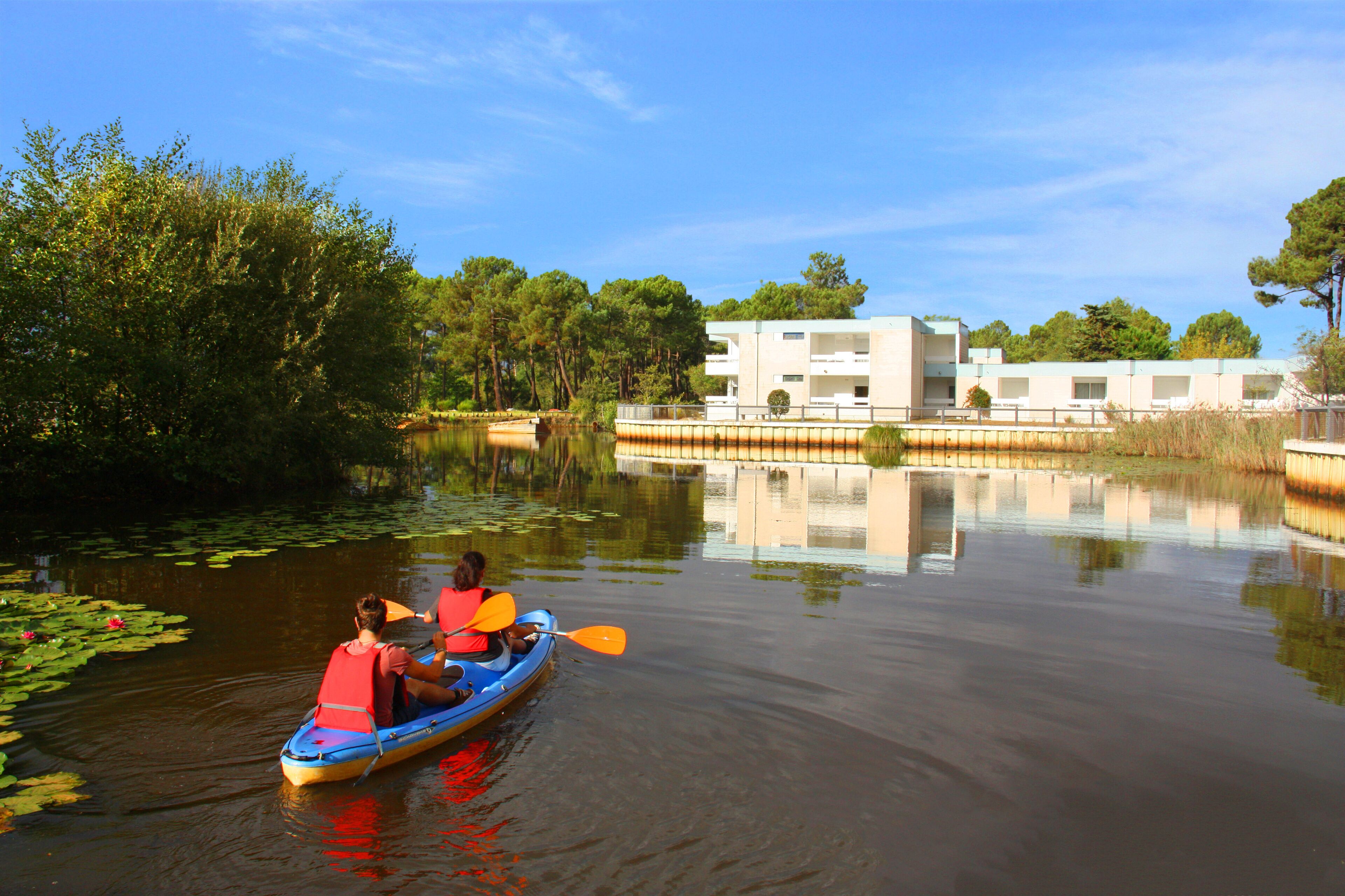Une rivière paisible, avec un canoë et une personne qui rame. On aperçoit, au loin, un bâtiment à la façade blanche.