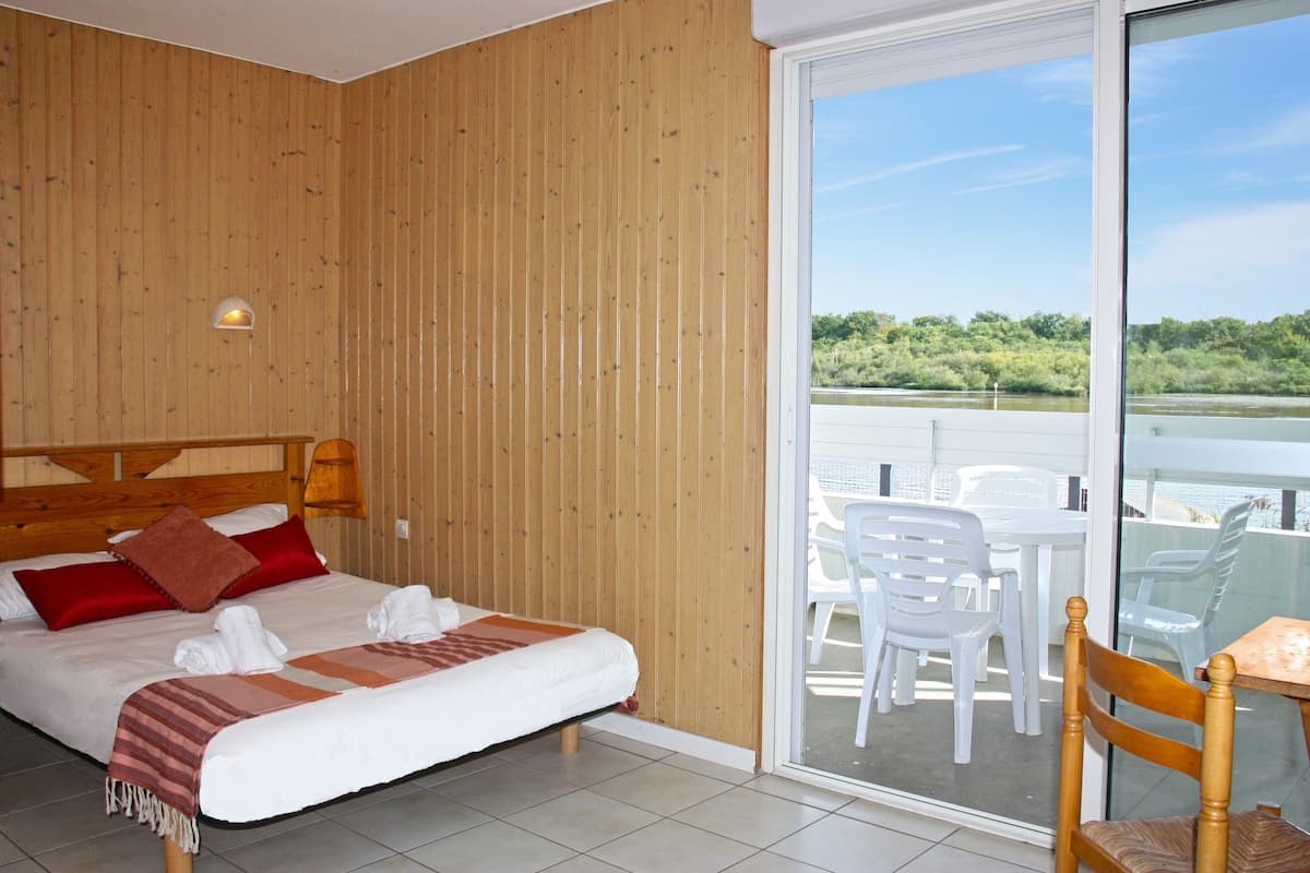 A hotel room with a wooden wall, a bed with red and brown pillows, a white blanket, and a view of a balcony with white chairs and a table.