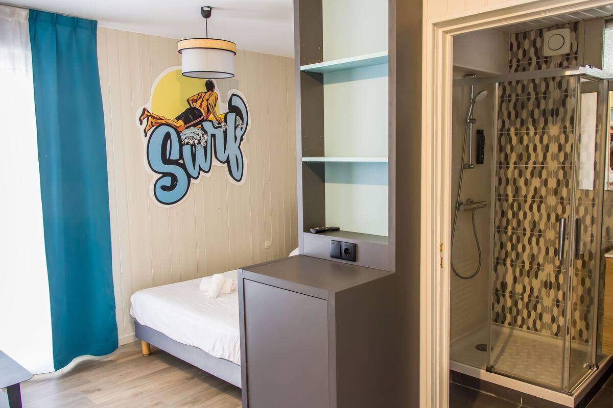 A modern hotel room with a bed, a small desk, a shower area, and a wall with a graphic.