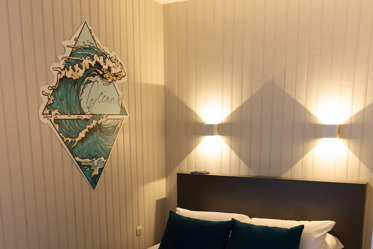 A neatly made bed with a dark green bedspread and white linens, a headboard with two wall-mounted lights, and a wall-mounted artwork depicting a wave.