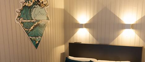 A neatly made bed with a dark green bedspread and white linens, a headboard with two wall-mounted lights, and a wall-mounted artwork depicting a wave.