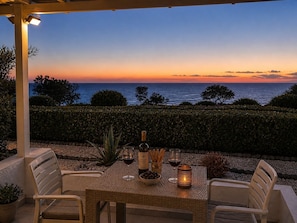 Un espace de restauration en plein air avec vue sur la mer au coucher du soleil, une bouteille de vin, des verres et une bougie sur une table.