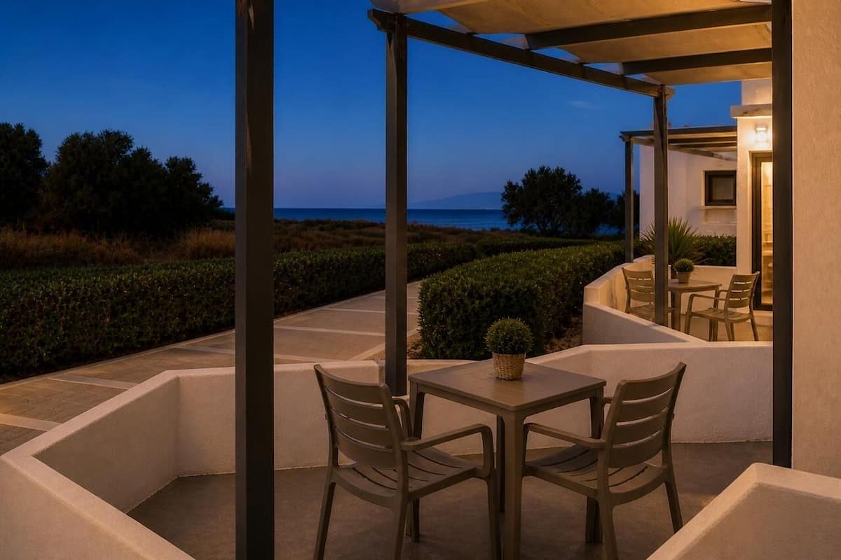 Un espacio al aire libre techado con mesas y sillas, una planta en maceta y vista al mar al atardecer.