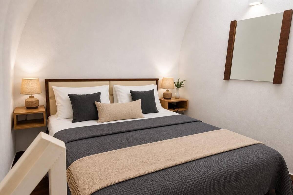 Una cama ordenadamente hecha con cabecera, dos mesitas de noche con lámparas y un espejo fijado a la pared.