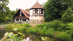 Una casa tradicional con entramado de madera y una torre, rodeada de vegetación exuberante y un estanque.