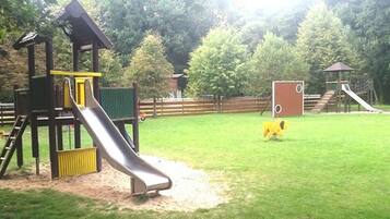 Explore the playground area with a wooden play structure featuring a slide and climbing area, set amidst a lush green field and shaded by trees.