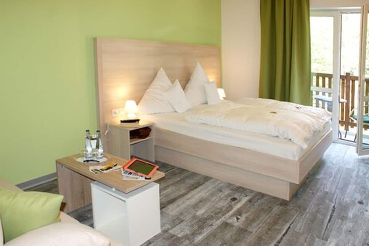 A bedroom with a wooden headboard, a bedside table with a lamp, a small bedside table with a tablet and water bottles, and a view of the outdoors through a door.