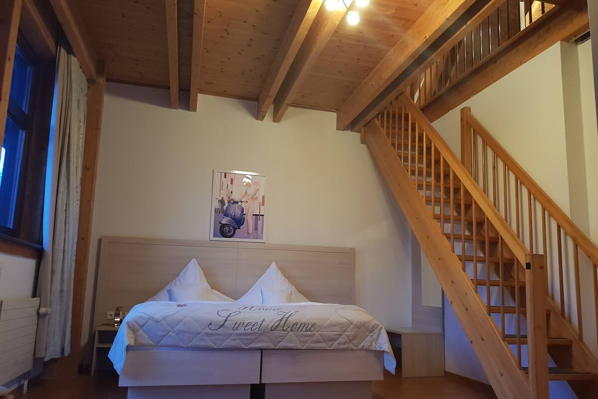 A bedroom with a wooden ceiling, a bed with a quilted headboard, a painting on the wall, and a wooden staircase.