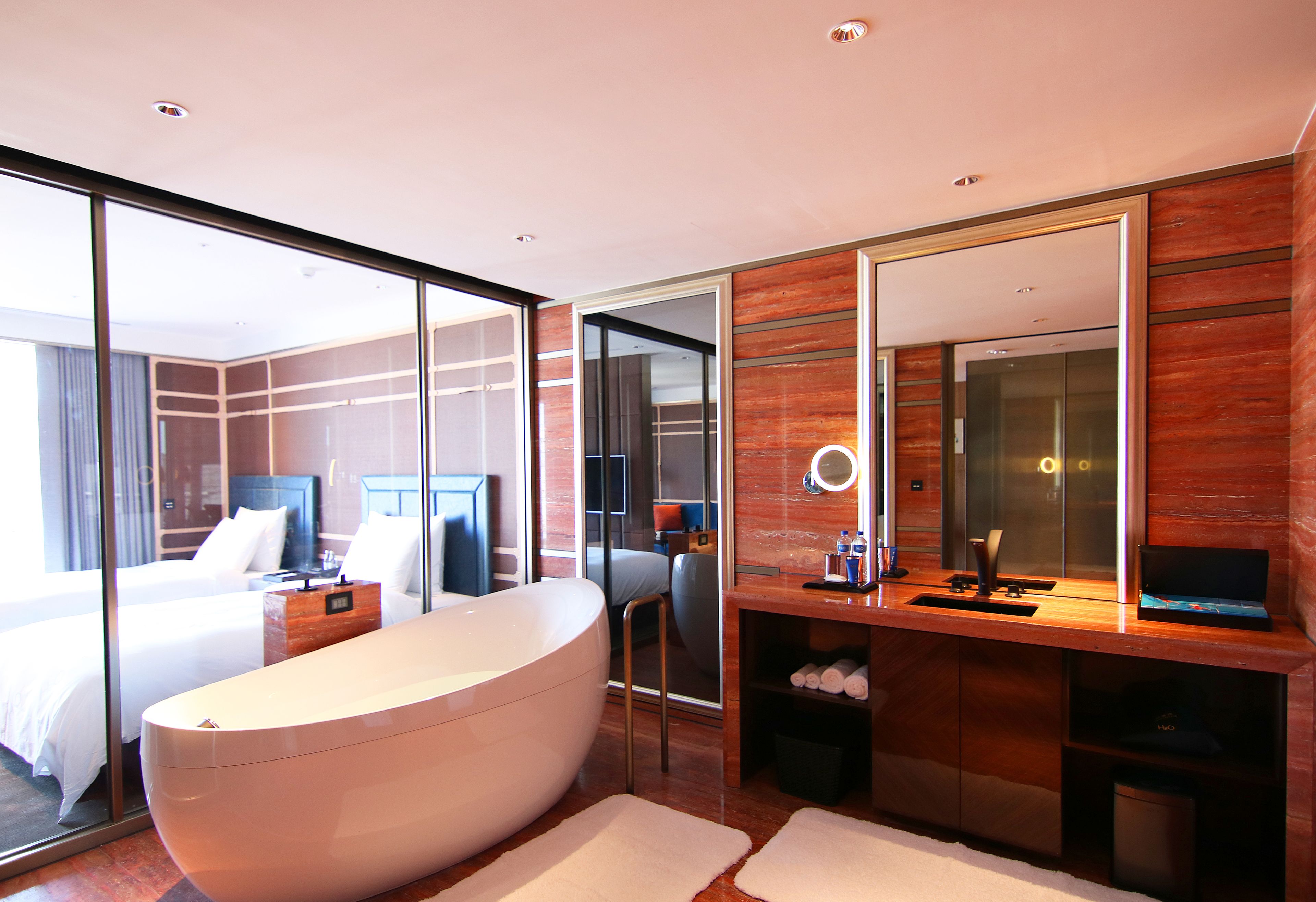 A modern hotel room with a large bathtub, a separate bedroom with a bed, a wooden vanity with a mirror, and a large window offering a view.