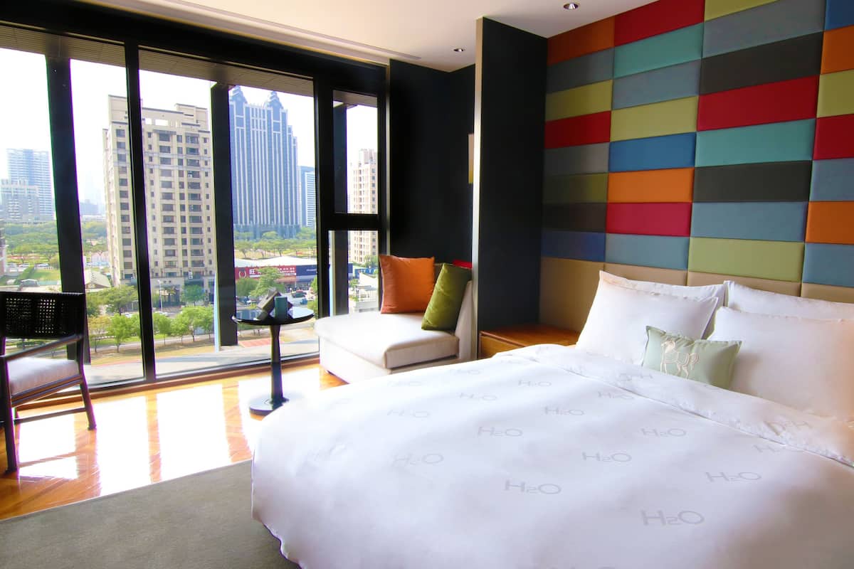 A hotel room with a large bed, a seating area with a chair and a small table, and a view of the city through large windows.