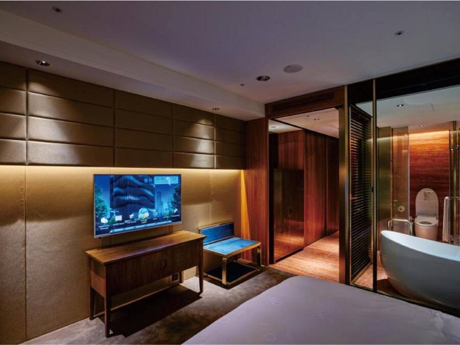 Une chambre d’hôtel moderne dotée d’un grand téléviseur à écran plat fixé au mur, d’un banc en bois, d’une douche en verre et d’une grande baignoire.