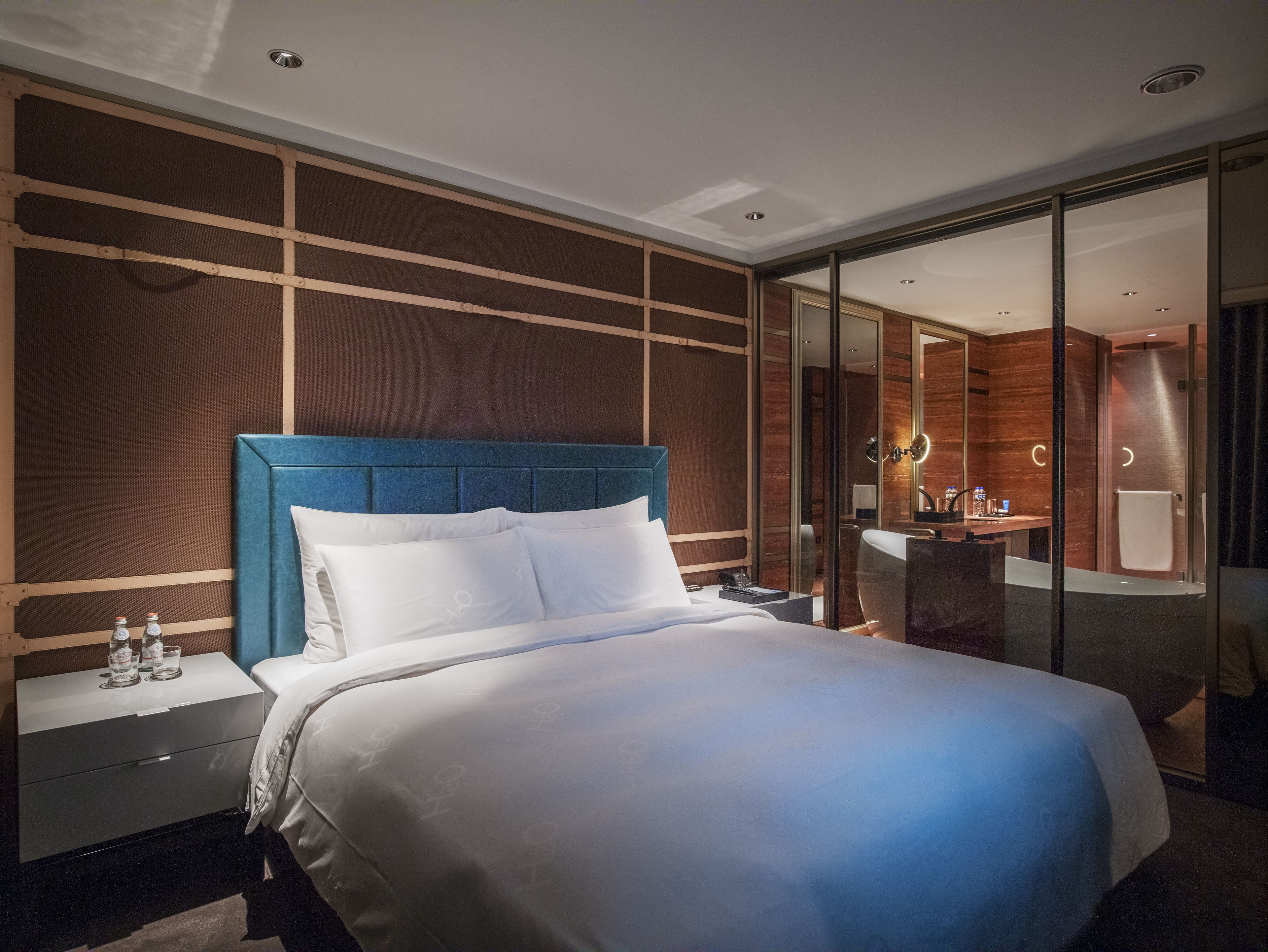 A modern hotel room with a large bed, a nightstand, a glass shower enclosure, and a bathroom with a mirror and sink.