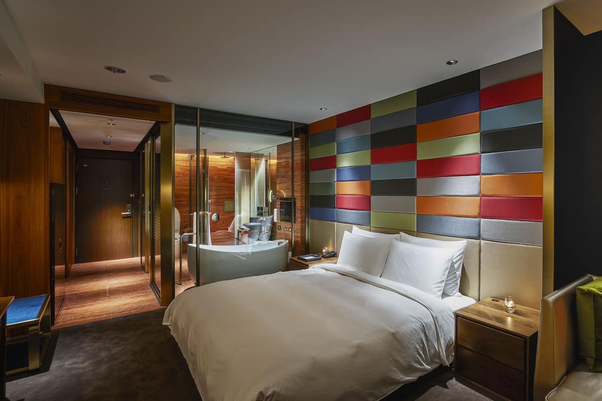 A modern hotel room with a large bed, a bedside table, a nightstand, a mirror, and a colorful wall panel.